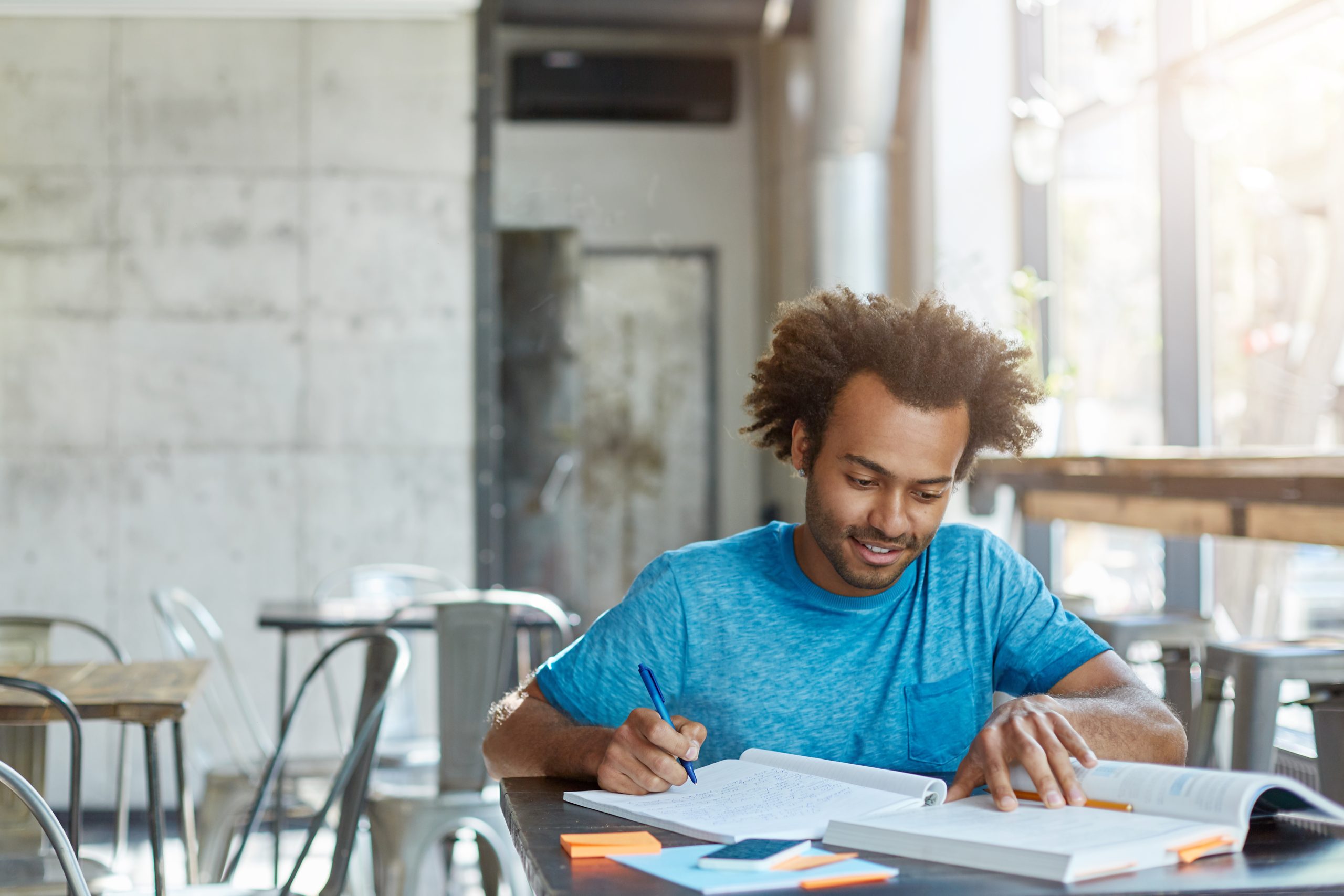 Hardworking excited African American A-student feeling happy after he solved complicated mathematical problem, smiling cheerfully and looking down in textbook while making notes in his copybook