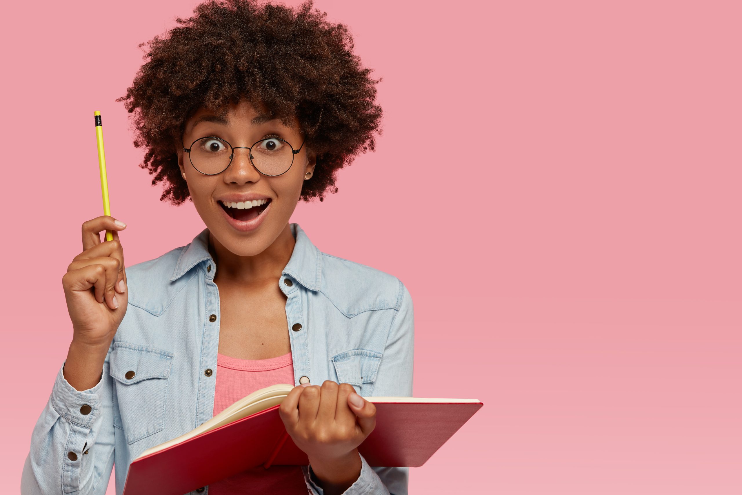 Indoor shot of cheerful dark skinned student keeps pencil in hand, feels happy as gets good idea for writing composition, holds notepad, works on article, looks joyfully isolated over pink studio wall